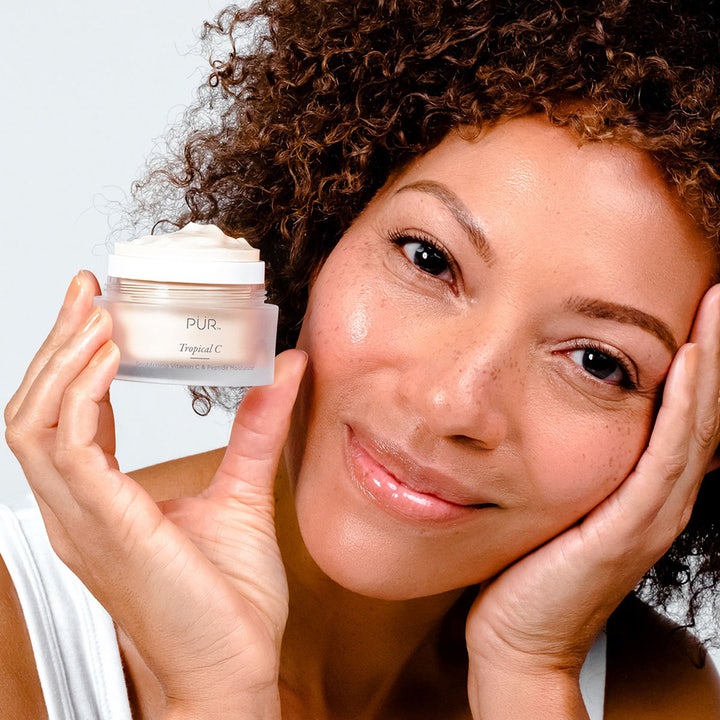 PÜR Tropical C Brightening Vitamin C & Peptide Moisturizer 50 ml dagcreme i åben krukke holdt af smilende kvinde.