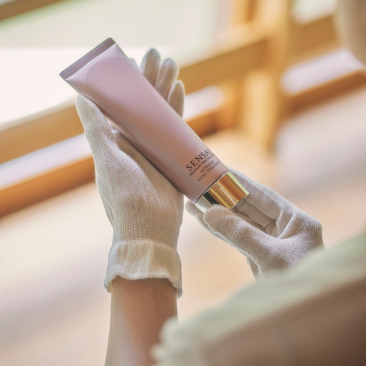 Sensai Cellular Performance Treatment Gloves 1 par i hvide bomuldshandsker der holder på en lyserød tube håndcreme