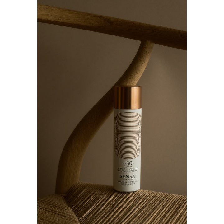 Sensai Silky Bronze Cooling Protective Suncare Spray SPF50 150 ml solcreme på en stilfuld flettet træstol.