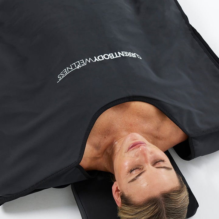 Currentbody Skin Sauna Blanket infrarødt saunatæppe til restitution viser en kvinde der slapper af i det sorte tæppe