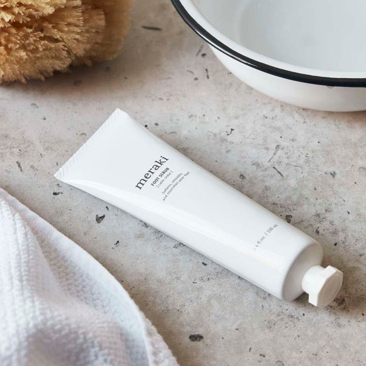 Meraki Footscrub Calm Cedar 100 ml eksfolierende fodcreme i hvid tube ved siden af et hvidt fad
