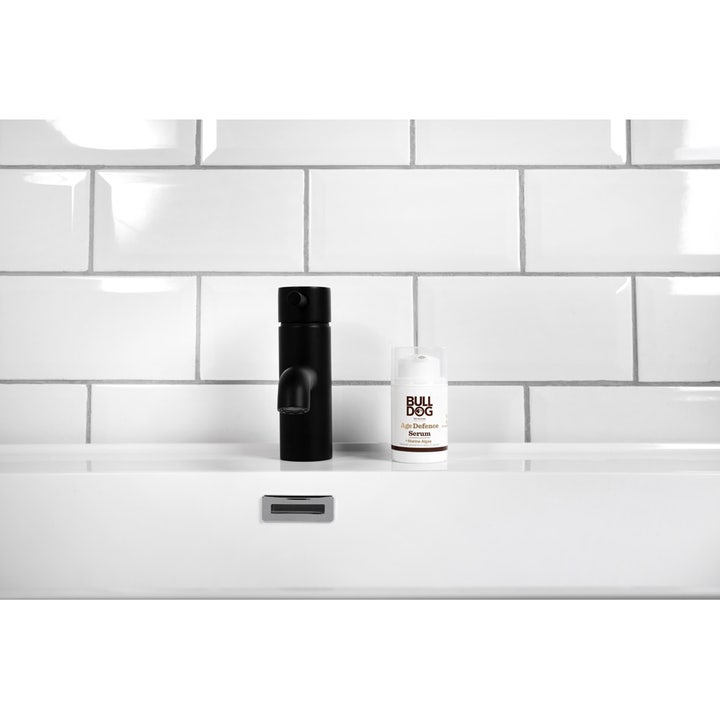Bulldog Age Defence Serum 50 ml modvirker rynker og ses i hvid pumpeflaske ved siden af en sort vandhane
