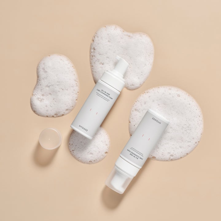 Hyggee All-In-One Care Cleansing Foam 150 ml ansigtsrens i hvide pumpeflasker omgivet af blødt, hvidt skum.