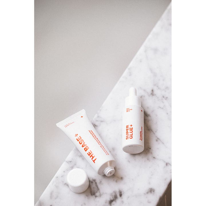 Swiss Clinic Super Glue+ 30 ml plejende serum i hvid pipetteflaske ved siden af The Base+ tube på hvid marmor.