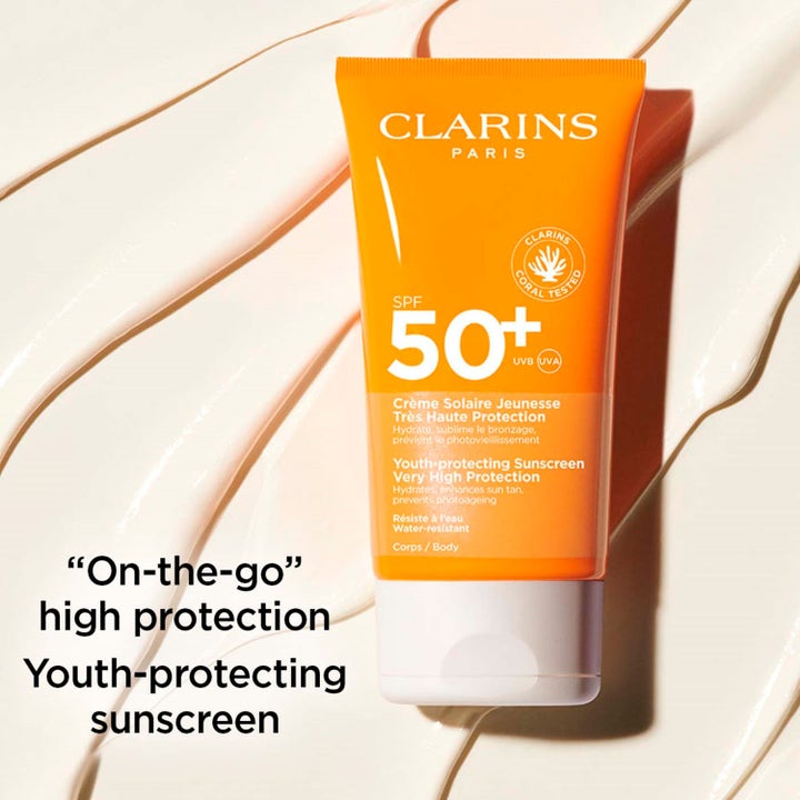 Clarins Youth-protecting Sunscreen 150 ml solcreme med SPF 50 i orange tube over cremet hvid tekstur.