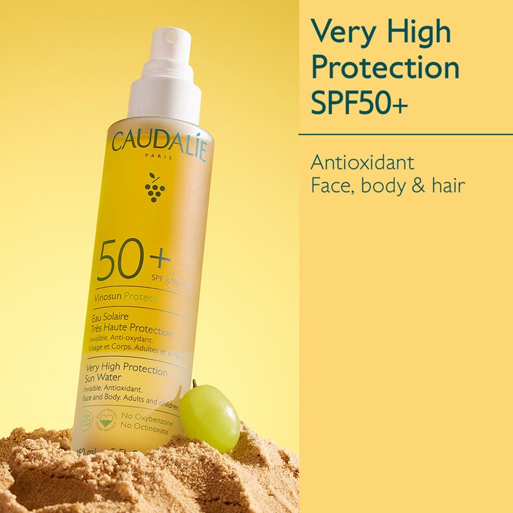 Caudalie Vinosun Very High Protection Water 150 ml solcreme SPF50+ i gul sprayflaske stående i sand med en drue