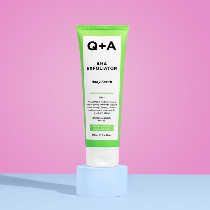 Q+A AHA Exfoliator Body Scrub 250 ml eksfolierende bodyscrub i en limegrøn tube stående på en lyseblå piedestal