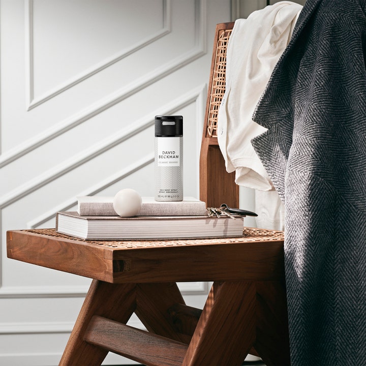 David Beckham Homme Deodorant Spray 150 ml giver friskhed i en hvid sprayflaske på bøger på en flettet træstol