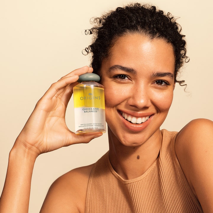 Origins Checks & Balances Milky Oil Cleanser 150 ml renseolie i to-faset flaske holdt af en smilende kvinde.
