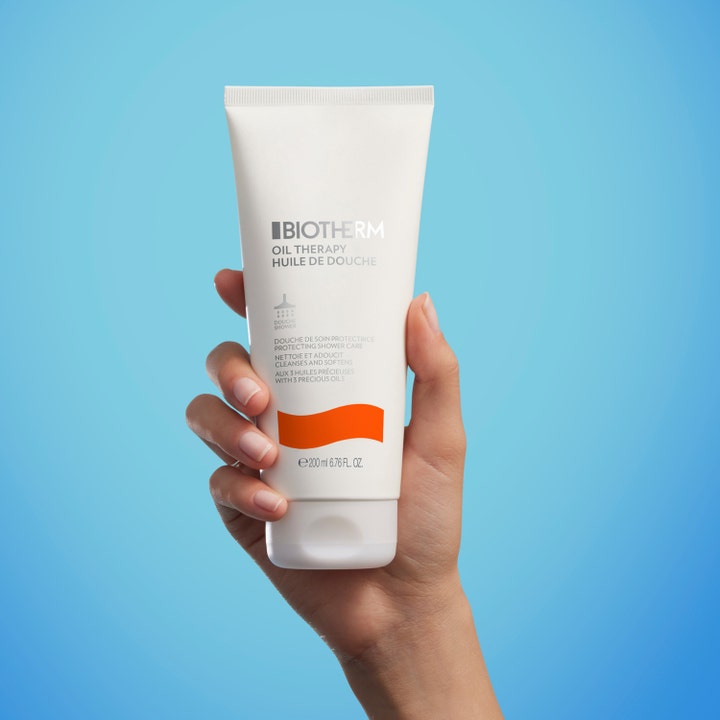 Biotherm Oil Therapy Baume Corps 200 ml plejende brusegel i hvid tube holdt af en hånd mod blå baggrund