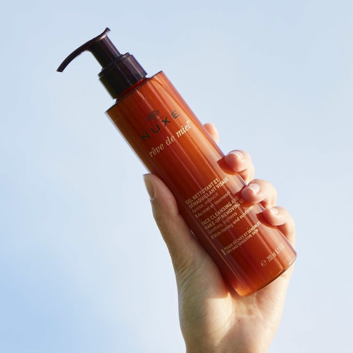Nuxe Rêve de Miel rensegel og makeupfjerner 200 ml beroligende ansigtsvask i ravfarvet flaske mod en blå himmel