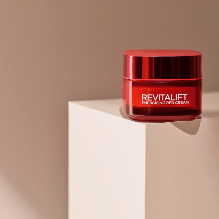 L'Oréal Paris Revitalift Ginseng Glow 50 ml opstrammende dagcreme i rød glaskrukke på en elegant beige sokkel.