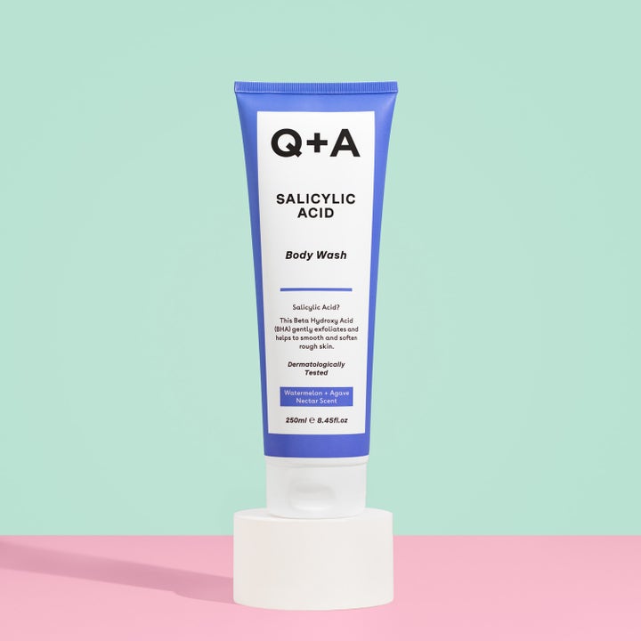 Q+A Salicylic Acid Body Wash 250 ml eksfolierende kropssæbe i en blå tube på en hvid piedestal mod pastelbaggrund