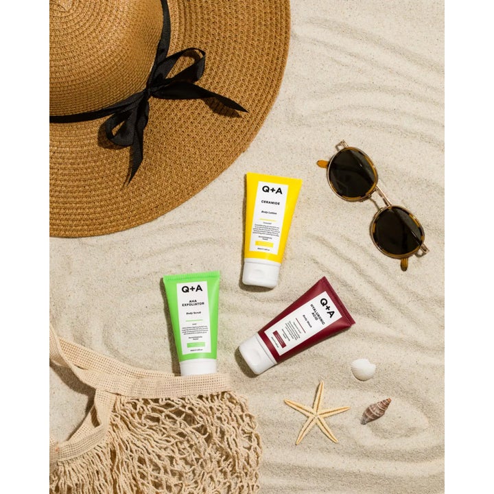 Q+A AHA Exfoliator Body Scrub 50 ml eksfolierende kropspleje i grøn tube på sand med solhat og solbriller
