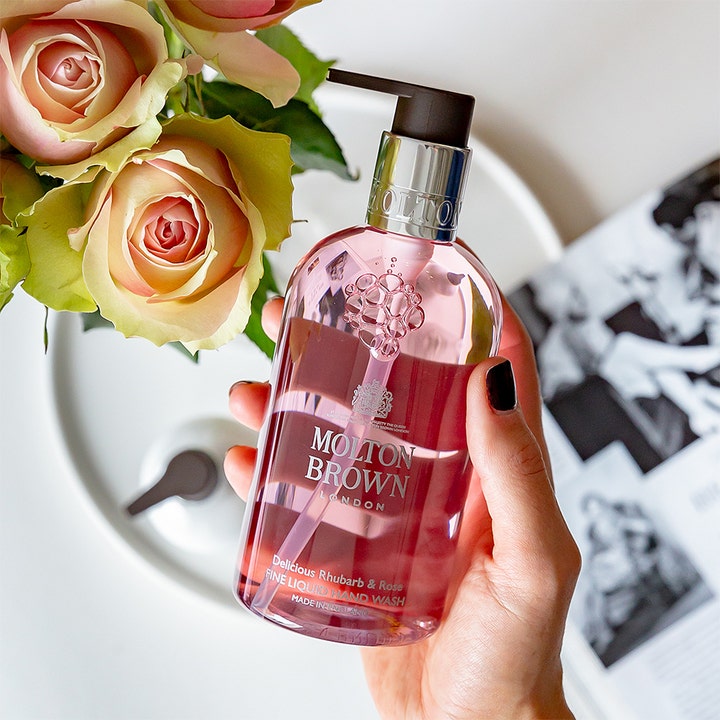Molton Brown Delicious Rhubarb & Rose 300 ml håndsæbe i klar lyserød flaske der holdes i hånden ved siden af friske roser