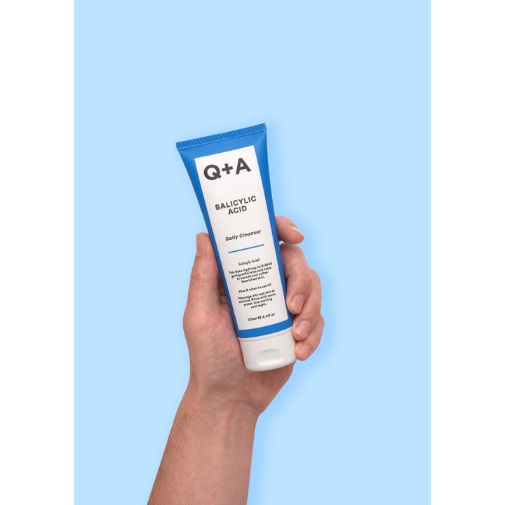 Q+A Salicylic Acid Cleanser 125 ml eksfolierende ansigtsrens i blå og hvid tube holdt i en hånd på blå baggrund.