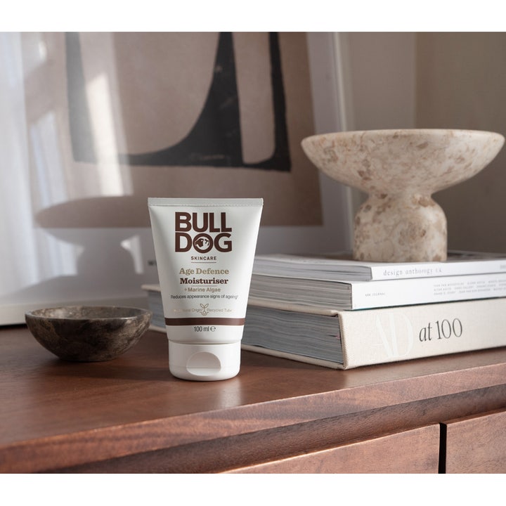 Bulldog Age Defence Moisturiser 100 ml fugtgivende ansigtscreme i hvid tube stående i et moderne interiør med bøger.