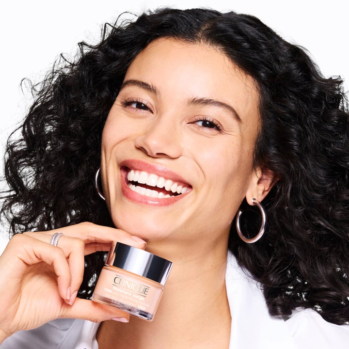 Clinique Moisture Surge Megastars fugtgivende ansigtscreme i krukke holdt af en smilende kvinde med krøller