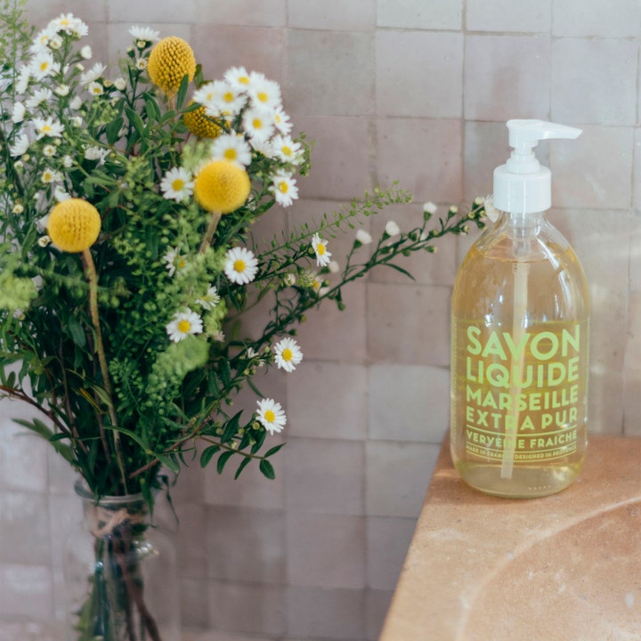 Compagnie de Provence Liquid Marseille Soap 495 ml håndsæbe i klar flaske ved en buket hvide og gule vilde blomster.