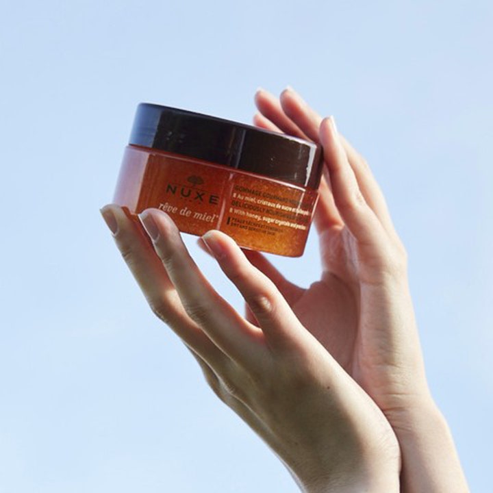 Nuxe Rêve de Miel Body Scrub 175 ml eksfolierende kropspleje i en krukke holdt af to hænder mod en blå himmel.