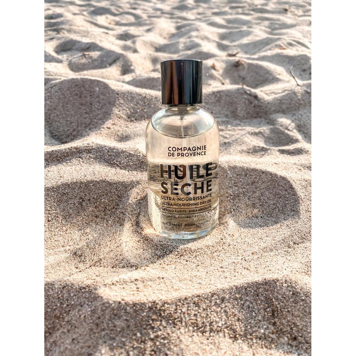 Compagnie de Provence Ultra Nourishing Dry Oil Shea Butter 100 ml nærende kropsolie i glasflaske på en solskinsstrand