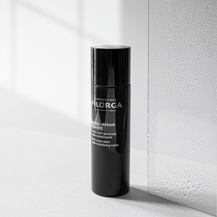Filorga Global-Repair Essence 150 ml revitaliserende essence i sort flaske ved siden af glas med vanddråber