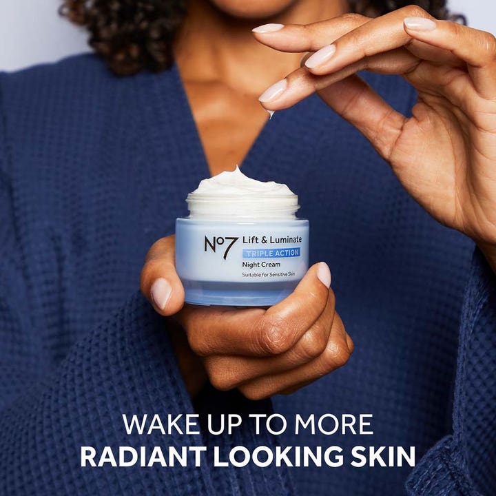 No7 Lift & Luminate Night Cream 50 ml giver glød i en blå krukke holdt af en person i mørkeblå morgenkåbe.