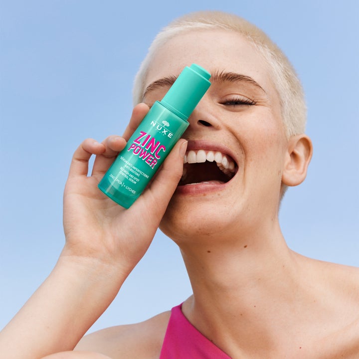 Nuxe Zinc Power Blemish And Spot Control Serum 30 ml mod urenheder holdes af en glad kvinde mod en klar blå himmel