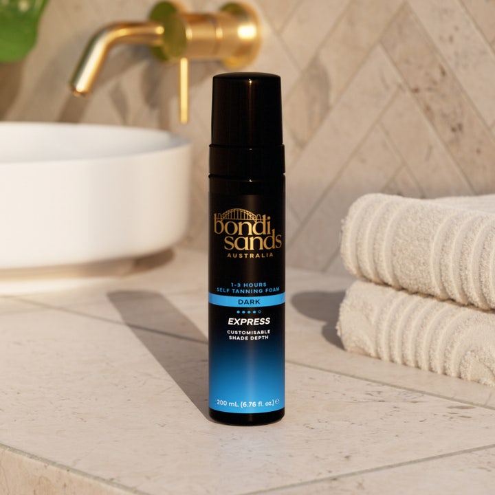 Bondi Sands Self Tan Foam 1 Hour Express 200 ml selvbruner i sort flaske ved siden af hvide håndklæder på et badeværelse.