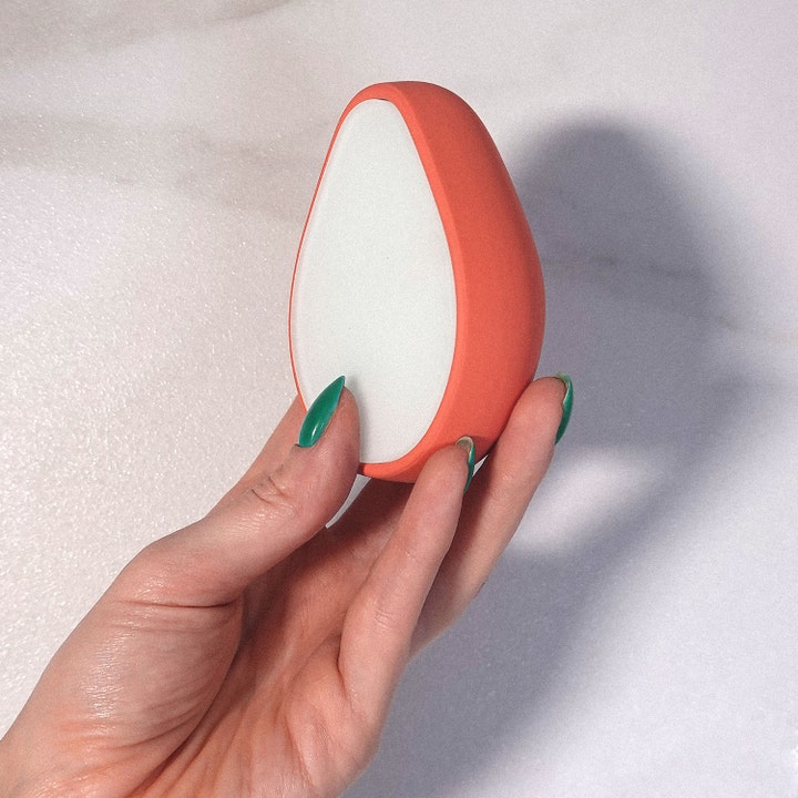 ANSWR Exfoliating Hair Removal Drop 1 stk. til hårfjerning i orange dråbeform holdt af en hånd med grønne negle.