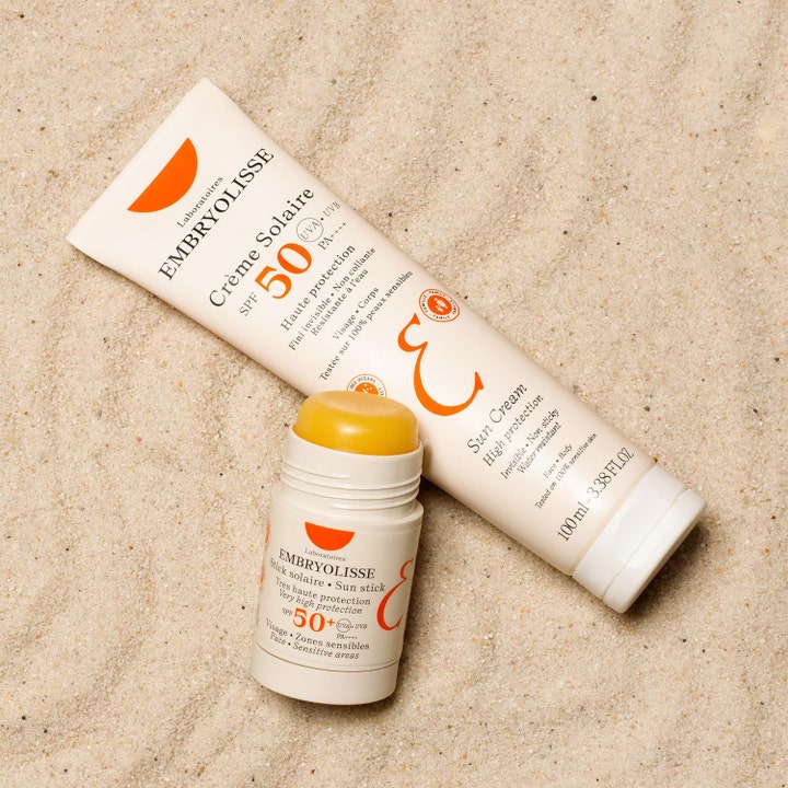 Embryolisse Sun Stick SPF50+ 15 g solstift med gul tekstur vist sammen med en tube solcreme på sand
